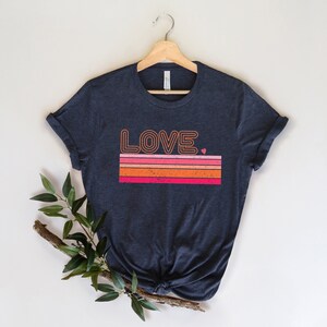 May include: A navy blue t-shirt with a retro-style graphic of the word "LOVE" in orange and pink with a small pink heart. The graphic is set against a background of orange, pink, and red stripes.