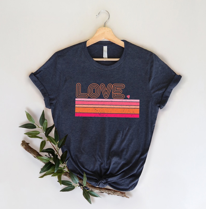 May include: A navy blue t-shirt with a retro-style graphic of the word "LOVE" in orange and pink with a small pink heart. The graphic is set against a background of orange, pink, and red stripes.