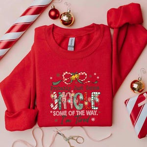 Pu&ograve; includere: Una felpa rossa con la scritta "Probably Just Gonna Jingle Some of the Way. I'm Tired" in un carattere festivo. L'immagine include decorazioni natalizie come palline, un regalo confezionato e alberi di Natale in miniatura.