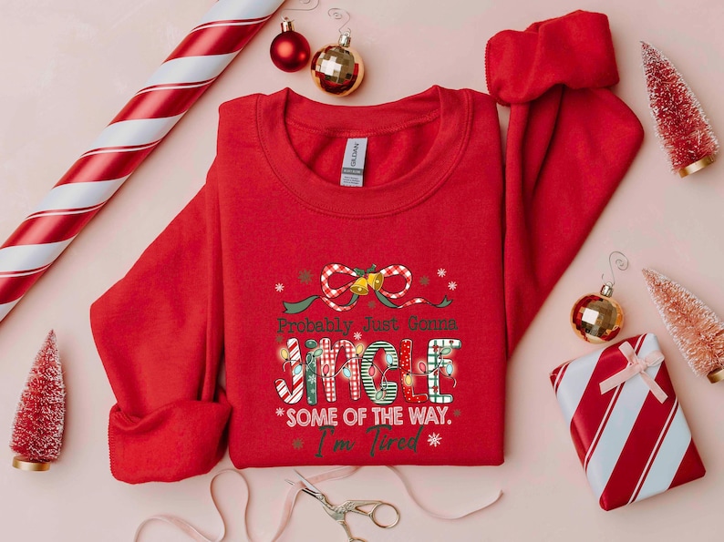Pu&ograve; includere: Una felpa rossa con la scritta "Probably Just Gonna Jingle Some of the Way. I'm Tired" in un carattere festivo. L'immagine include decorazioni natalizie come palline, un regalo confezionato e alberi di Natale in miniatura.