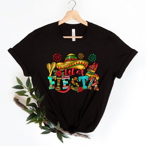 Puede incluir: Camiseta negra con un dise&ntilde;o gr&aacute;fico colorido que incluye un sombrero, maracas y el texto "Let's Fiesta!"
