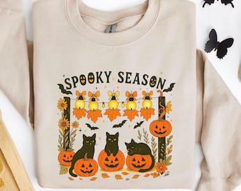 May include: Beige sweatshirt with a Halloween-themed graphic. The design features black cats sitting on carved pumpkins, string lights, fall leaves, and the words "Spooky Season". The design is framed by sunflowers and wooden posts.