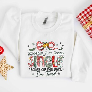 Pu&ograve; includere: Felpa bianca con la scritta "Probably Just Gonna Jingle Some of the Way. I'm Tired." La parola "Jingle" &egrave; decorata con luci natalizie e motivi a quadri. Un fiocco rosso e verde e fiocchi di neve sono sopra il testo. A tema natalizio.