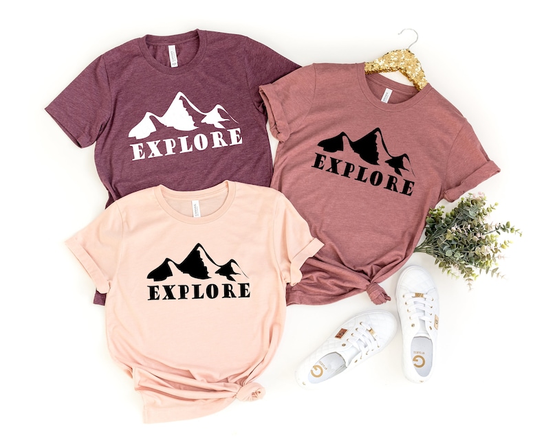 May include: Three t-shirts with a mountain range graphic and the word "EXPLORE" printed on them. The shirts are in different colors: maroon, peach, and light pink.