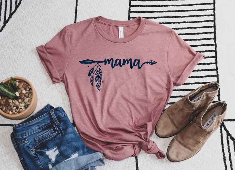 May include: A mauve t-shirt with a navy blue arrow and feathers design. The text "mama" is written in a cursive font on the shirt.