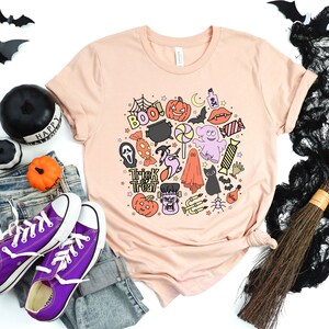 May include: A peach-colored t-shirt with a Halloween-themed design featuring a variety of icons, including a ghost, a pumpkin, a cauldron, a spider, and the words "Trick or Treat" and "Boo!"