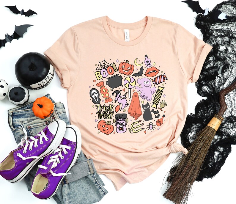 May include: A peach-colored t-shirt with a Halloween-themed design featuring a variety of icons, including a ghost, a pumpkin, a cauldron, a spider, and the words "Trick or Treat" and "Boo!"