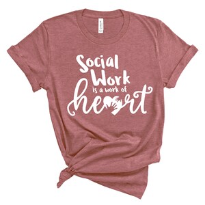 May include: A maroon t-shirt with the text "Social Work is a work of heart" in white lettering. The heart graphic features two hands holding each other.
