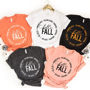 May include: Four different colored t-shirts with a white circle graphic that says "Hello Fall" with fall-themed text around the circle. The shirts are orange, white, black, and pink.