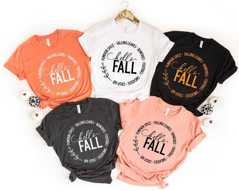 May include: Four different colored t-shirts with a white circle graphic that says "Hello Fall" with fall-themed text around the circle. The shirts are orange, white, black, and pink.
