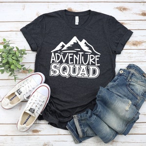 Puede incluir: Una camiseta gris oscuro con un gr&aacute;fico blanco de monta&ntilde;as y el texto "Adventure Squad". La camiseta est&aacute; colocada sobre una superficie de madera blanca con un par de zapatos de lona blancos y unos vaqueros azules.