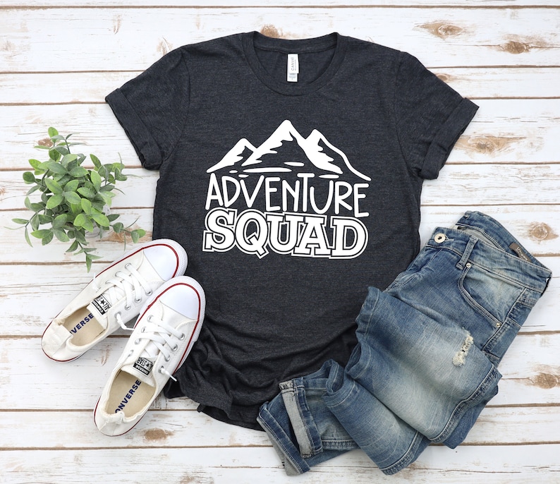 Puede incluir: Una camiseta gris oscuro con un gr&aacute;fico blanco de monta&ntilde;as y el texto "Adventure Squad". La camiseta est&aacute; colocada sobre una superficie de madera blanca con un par de zapatos de lona blancos y unos vaqueros azules.
