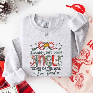 Pu&ograve; includere: Una felpa grigio chiaro con la scritta "Probably Just Gonna Jingle Some of the Way. I'm Tired" in un design festivo e colorato. Il design include un fiocco rosso e bianco, luci natalizie e stelle. &Egrave; visibile un sacchetto regalo a tema natalizio.