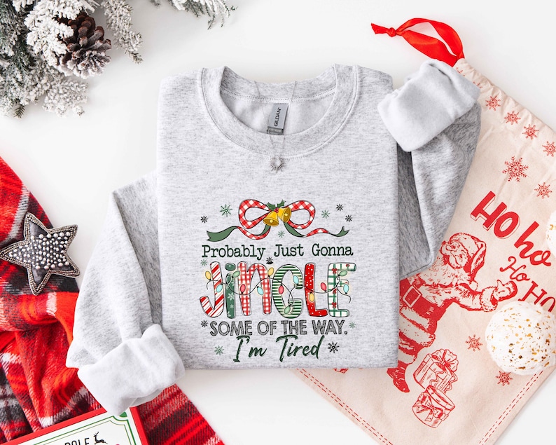 Pu&ograve; includere: Una felpa grigio chiaro con la scritta "Probably Just Gonna Jingle Some of the Way. I'm Tired" in un design festivo e colorato. Il design include un fiocco rosso e bianco, luci natalizie e stelle. &Egrave; visibile un sacchetto regalo a tema natalizio.