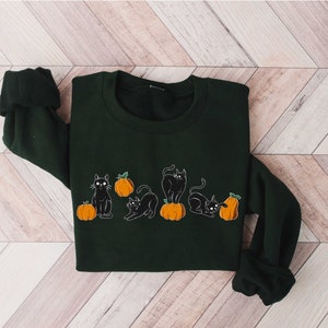 Puede incluir: Una sudadera verde oscuro con un dise&ntilde;o de gato negro y calabaza. Los gatos est&aacute;n en varias poses, y las calabazas son de color naranja con tallos verdes.