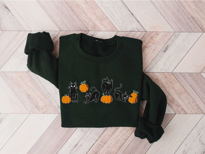 Puede incluir: Una sudadera verde oscuro con un dise&ntilde;o de gato negro y calabaza. Los gatos est&aacute;n en varias poses, y las calabazas son de color naranja con tallos verdes.