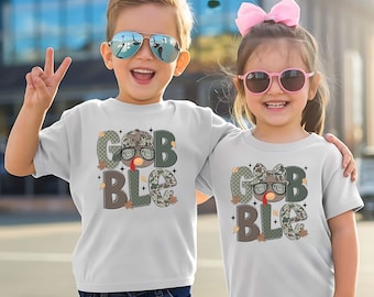 May include: Two children wearing white t-shirts with a "Gobble" design. The design features a turkey wearing sunglasses and a camouflage bow tie or hat. The word "Gobble" is in a mix of camouflage and brown letters. The children are smiling and wearing sunglasses.