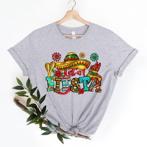 Puede incluir: Camiseta gris con un dise&ntilde;o gr&aacute;fico colorido que dice "Let's Fiesta". El dise&ntilde;o incluye un sombrero, maracas y flores.