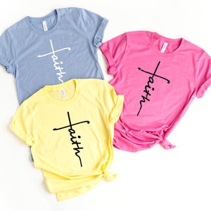 Op de afbeelding: Drie T-shirts met het woord "geloof" verticaal geschreven met een kruis voor het woord. De shirts zijn in blauw, geel en roze.