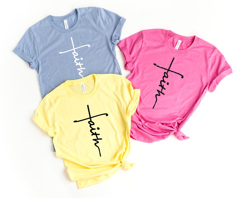 Op de afbeelding: Drie T-shirts met het woord "geloof" verticaal geschreven met een kruis voor het woord. De shirts zijn in blauw, geel en roze.