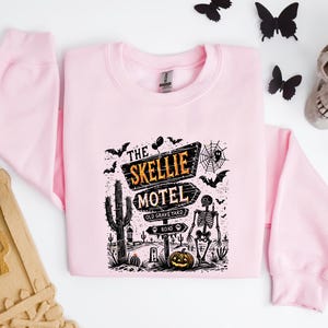 May include: A light pink sweatshirt with a graphic featuring a Halloween-themed design. The graphic includes the text "The Skellie Motel" with a skeleton, cactus, and pumpkin. Black bats and a spiderweb are also included in the design.