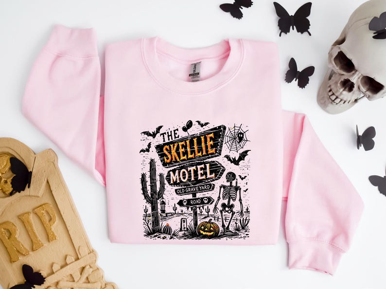 May include: A light pink sweatshirt with a graphic featuring a Halloween-themed design. The graphic includes the text "The Skellie Motel" with a skeleton, cactus, and pumpkin. Black bats and a spiderweb are also included in the design.