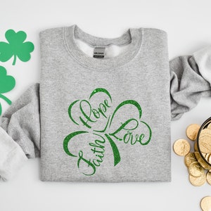 Puede incluir: Una sudadera gris con un dise&ntilde;o de tr&eacute;bol verde con purpurina que dice "Fe, Esperanza, Amor".