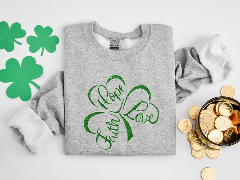 Puede incluir: Una sudadera gris con un dise&ntilde;o de tr&eacute;bol verde con purpurina que dice "Fe, Esperanza, Amor".
