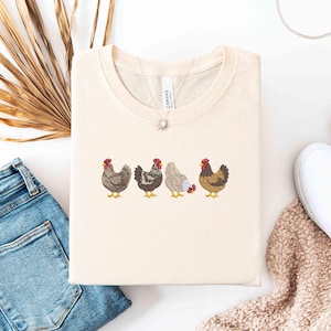 May include: Cream-colored t-shirt featuring an embroidered design of four chickens in a row. The chickens are in various colors, including brown, gray, and white, with red combs and yellow feet. The shirt is folded neatly, with a necklace visible.