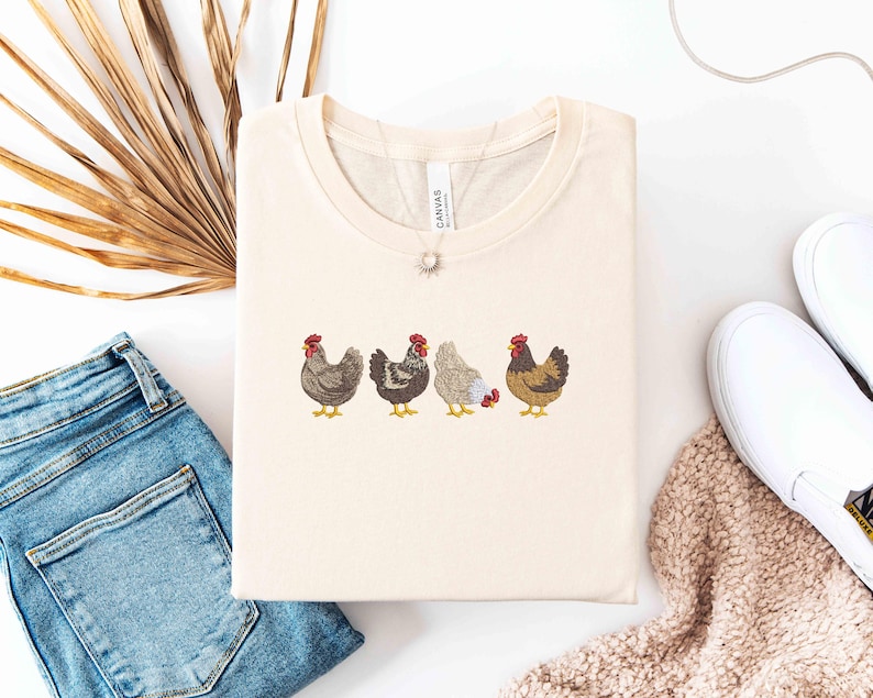 May include: Cream-colored t-shirt featuring an embroidered design of four chickens in a row. The chickens are in various colors, including brown, gray, and white, with red combs and yellow feet. The shirt is folded neatly, with a necklace visible.