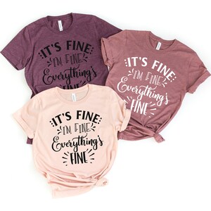 Peut inclure: Trois t-shirts avec le texte "It's fine: I'm fine. Everything's fine." en &eacute;criture cursive noire sur un fond rose et mauve.