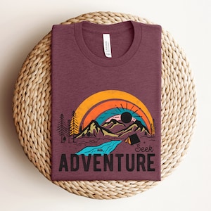 May include: Burgundy t-shirt with a graphic of a mountain landscape, rainbow, and the words "Seek Adventure." The design includes a river, trees, and a tent. The shirt is folded on a woven, circular surface.