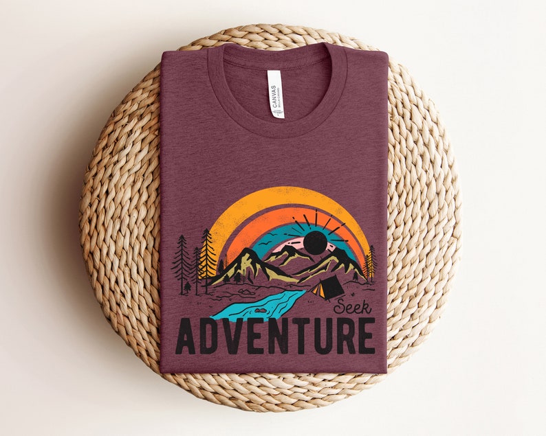 May include: Burgundy t-shirt with a graphic of a mountain landscape, rainbow, and the words "Seek Adventure." The design includes a river, trees, and a tent. The shirt is folded on a woven, circular surface.