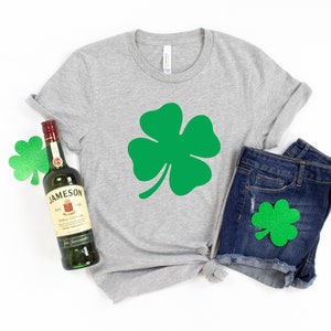 K&ouml;nnte beinhalten: Ein graues T-Shirt mit einem gr&uuml;nen vierbl&auml;ttrigen Kleeblatt-Motiv, eine Flasche Jameson Irish Whiskey und eine blaue Jeans-Shorts mit einem gr&uuml;nen vierbl&auml;ttrigen Kleeblatt-Aufn&auml;her.
