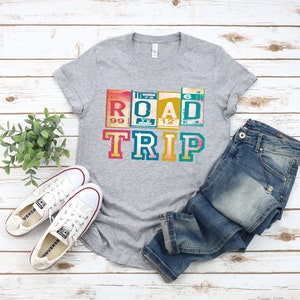 May include: A gray t-shirt with a vintage-style graphic that says "ROAD TRIP" in colorful letters. The letters are made to look like old road signs. The shirt is laid out on a white wooden surface with a pair of white sneakers and blue jeans.