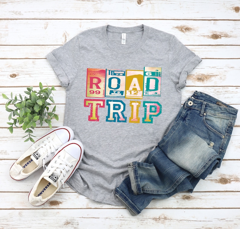 May include: A gray t-shirt with a vintage-style graphic that says "ROAD TRIP" in colorful letters. The letters are made to look like old road signs. The shirt is laid out on a white wooden surface with a pair of white sneakers and blue jeans.