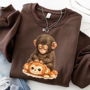 May include: A brown sweatshirt featuring a graphic of a baby monkey with closed eyes, gently touching a plush toy monkey. A silver necklace with a small charm is visible. The sweatshirt has a crew neck and long sleeves.