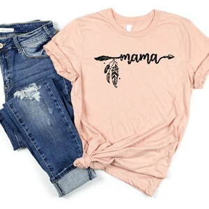 May include: A peach-colored t-shirt with a black arrow and feathers design and the word "mama" written in a script font.