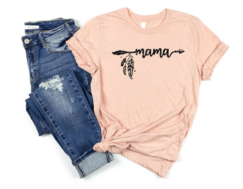 May include: A peach-colored t-shirt with a black arrow and feathers design and the word "mama" written in a script font.
