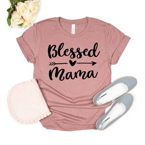 May include: A dusty rose t-shirt with the text "Blessed Mama" in black lettering. The text includes a small heart and arrow graphic.