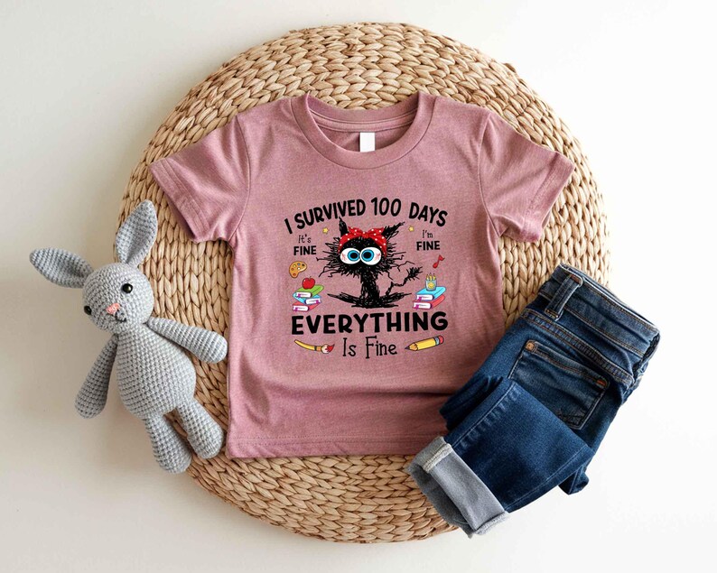 May include: A mauve short-sleeved t-shirt with the text "I Survived 100 Days Everything Is Fine" and a cartoon cat graphic. The shirt is paired with blue denim jeans. A gray stuffed bunny is also in the image.
