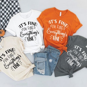 Peut inclure: Quatre t-shirts avec le texte "It's fine: I'm fine. Everything's fine." en lettres noires sur un t-shirt blanc, beige, orange et gris. Les t-shirts sont dispos&eacute;s sur une surface gris clair avec un jean bleu au centre.