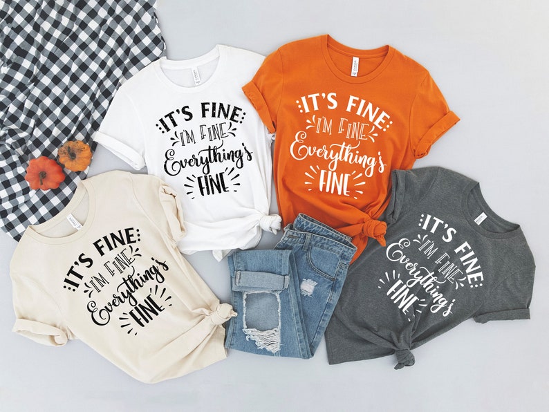 Peut inclure: Quatre t-shirts avec le texte "It's fine: I'm fine. Everything's fine." en lettres noires sur un t-shirt blanc, beige, orange et gris. Les t-shirts sont dispos&eacute;s sur une surface gris clair avec un jean bleu au centre.