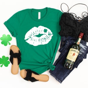 May include: A green t-shirt with a white graphic of a kiss with a shamrock on it. The shirt is laid out on a white background with a pair of black sandals, blue jeans, a black lace top, and a bottle of Jameson Irish Whiskey.