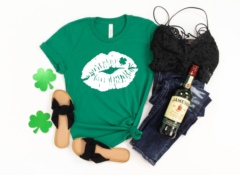 May include: A green t-shirt with a white graphic of a kiss with a shamrock on it. The shirt is laid out on a white background with a pair of black sandals, blue jeans, a black lace top, and a bottle of Jameson Irish Whiskey.