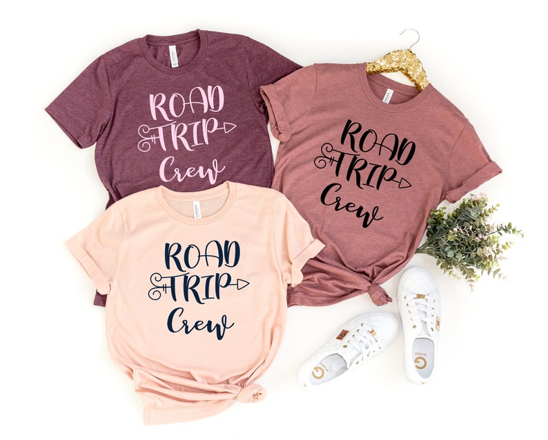 May include: Three t-shirts with the text "Road Trip Crew" in different colors. The shirts are maroon, peach, and a darker maroon. The text is in black and pink on the maroon shirts and in black and navy blue on the peach shirt.
