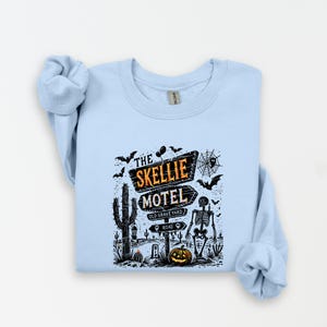 May include: Light blue sweatshirt featuring a Halloween-themed graphic. The design includes a sign that reads "The Skellie Motel," a skeleton, bats, a cactus, and a jack-o'-lantern. The graphic is in black, orange, and white.