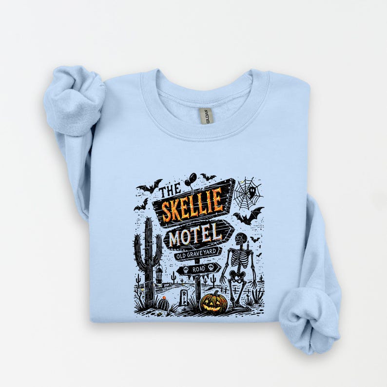 May include: Light blue sweatshirt featuring a Halloween-themed graphic. The design includes a sign that reads "The Skellie Motel," a skeleton, bats, a cactus, and a jack-o'-lantern. The graphic is in black, orange, and white.