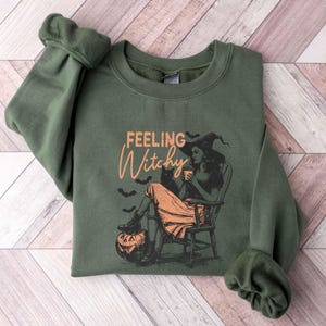 May include: Olive green sweatshirt with the text "Feeling Witchy" in orange script. The graphic features a witch in a hat, sitting in a chair with a black cat, a pumpkin, and bats. A cozy, Halloween-themed garment.