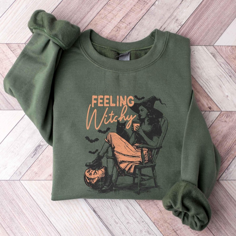 May include: Olive green sweatshirt with the text "Feeling Witchy" in orange script. The graphic features a witch in a hat, sitting in a chair with a black cat, a pumpkin, and bats. A cozy, Halloween-themed garment.
